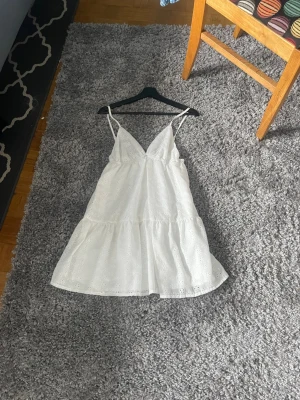 Vit kort klänning med spetsdetaljer - Söt vit klänning med tunna axelband och v-ringning. Klänningen har spetsdetaljer och broderade blommor, samt en volang nedtill som ger en luftig känsla. Perfekt för varma dagar och sommarens alla tillfällen.