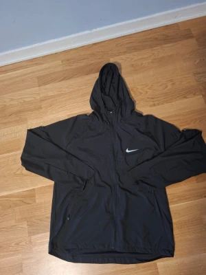 Svart Nike vindjacka  - Snygg svart vindjacka från Nike med hel dragkedja framtill och justerbar huva. Jackan har två sidofickor med dragkedja och den klassiska vita Nike-loggan på bröstet. Tillverkad i lätt och vindtätt material, perfekt för blåsiga dagar.