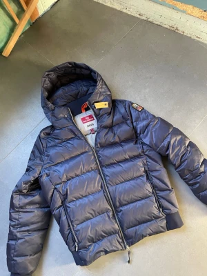 Parajumper jacka - Snygg mörkblå dunjacka från Parajumpers med huva och dragkedja framtill.                                   Lite liten i storleken passar 173-185.                    Den är i väldigt bra skick men den är så billig för dragkedjan gått sönder,     går fortfarande å stänga om man drar upp å ner med den som är kvar men den andra har gått av.                           Tveka inte på å skriva om du vill ge ett bud eller fråga nått!