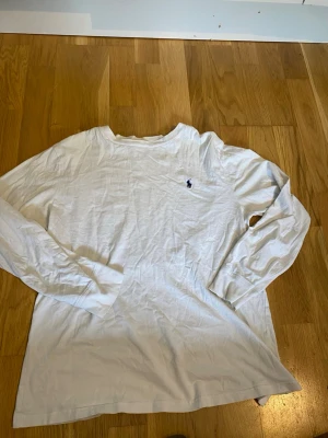 Vit långärmad tröja från Ralph Lauren - Klassisk vit långärmad tröja från Ralph Lauren med rund halsringning och den ikoniska lilla broderade loggan på bröstet. Tillverkad i mjuk bomull som känns skön mot huden. Perfekt att matcha med jeans eller shorts för en clean look.