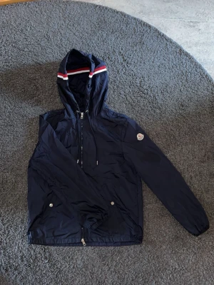 Mörkblå vindjacka från Moncler. - Snygg mörkblå vindjacka från Moncler med huva och dragkedja. Perfekt för dig som vill klä sig stilligt men även svalt inför våren och sommaren.