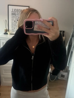 Svart stickad ziptröja - Mysig svart stickad tröja med dragkedja framtill och långa ärmar. Tröjan har en croppad passform och ribbade muddar vid ärmslut och nederkant. Perfekt att slänga på sig över ett linne eller t-shirt.