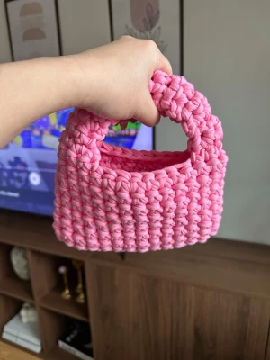 Handvirkad rosa handväska - Supersöt handväska i chunky virkat material och stark rosa färg. Väskan har en kompakt, rektangulär form med ett matchande handtag. Perfekt accessoar för dig som vill sticka ut med en unik och trendig look.