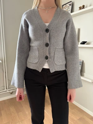 Grå kofta med stora knappar - Säljer en stilren grå kofta med v-ringning och tre stora svarta knappar framtill. Koftan har två fickor fram och är tillverkad i ett mjukt stickat material. Ärmarna är raka och något vida för en avslappnad look.
