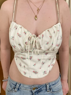 Vit blommig korsettopp med snörning - Supersöt vit korsettopp med små rosa blommor och snörning både fram och bak. Toppen har smala axelband och en figurnära passform som ger en snygg siluett. Perfekt för dig som vill ha en romantisk och trendig look.