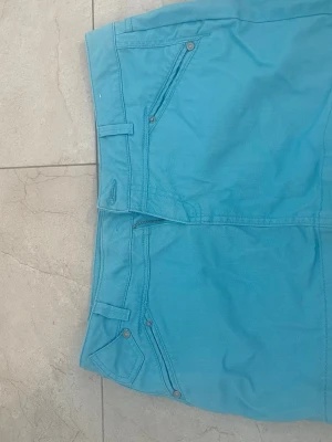 Turkos kortkjol med fickor - Säljer en turkos kortkjol med klassiska jeansfickor fram och bak samt bälteshällor. Kjolen har dragkedja framtill och är gjord i ett bomullsliknande material. Perfekt för dig som vill ha en färgglad och enkel kjol till sommaren. Knappen skanas