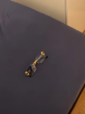 Blå rektangulära solglasögon guld - Säljer ett par trendiga solglasögon med rektangulära blå glas och guldfärgade metallbågar. Glasögonen har minimalistisk design utan synlig ram runt glasen och svarta skalmar. Perfekt accessoar för dig som vill sticka ut.