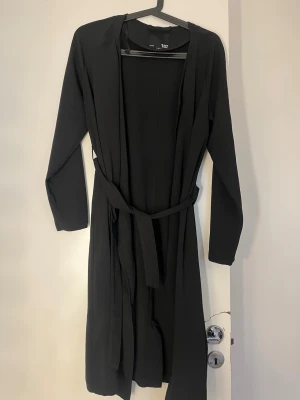 Svart morgonrock med skärp H&M - En stilren svart morgonrock från H&M med långa ärmar och matchande midjebälte. Morgonrocken har en enkel och klassisk design, perfekt för att chilla hemma. Tillverkad i ett mjukt och lätt material som känns skönt mot huden.