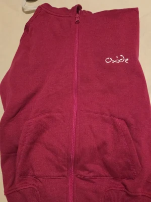 Vinröd hoodie med dragkedja från Oxide - Snygg vinröd hoodie från Oxide med dragkedja framtill och två stora fickor. Broderad logga på bröstet och klassisk huva. Perfekt för en avslappnad och trendig stil. Tillverkad i mjukt material som känns skönt mot huden.