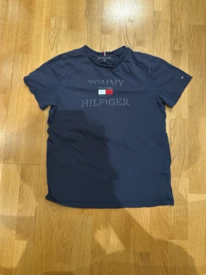 Mörkblå Tommy Hilfiger t-shirt - Snygg mörkblå t-shirt från Tommy Hilfiger med klassisk logga och flagga på bröstet. Rund halsringning och korta ärmar. Tillverkad i mjuk bomull för skön känsla. Perfekt för dig som gillar stilrena och tidlösa plagg med en sportig touch.