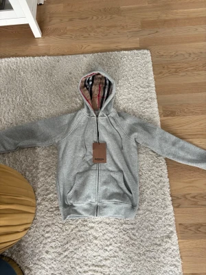 Grå hoodie från Burberry - Säljer en stilren grå hoodie från Burberry med dragkedja framtill och klassiskt randigt foder i huvan. Hoodien har fickor på sidorna och ribbade muddar vid ärmslut och nederkant. Perfekt för en chill och trendig look.
