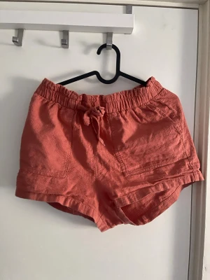 Koralrosa shorts med snörning - Säljer ett par korallrosa shorts med snörning i midjan och avslappnad passform. Shortsen har bred resår i midjan och stora fickor framtill. Perfekta för varma dagar och tillverkade i mjuk bomull.