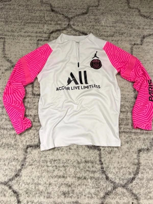 PSG träningsset Jordan x Paris  - Säljer ett fett PSG träningsset från Jordan x Paris Saint-Germain. Tröjan är vit med rosa och svarta detaljer, halv dragkedja och coola mönstrade rosa ärmar. Byxorna är svarta med rosa detaljer och Paris-tryck på benet. Perfekt för fotbollsträning eller chill.