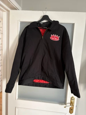 Svart vindjacka från DePalma - Svart vindjacka från DePalma med röd broderad logga på bröstet och stort broderi på ryggen. Jackan har dragkedja framtill, två sidofickor och rött innerfoder. Materialet är troligtvis polyester eller nylon, perfekt för en streetwear-look.