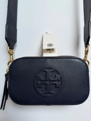 Mörkblå Miller mini crossbody från Tory Burch - Snygg mörkblå Miller mini crossbody från Tory Burch i präglat läder med det klassiska logotypmönstret framtill. Väskan har en bred axelrem med guldfärgade metalldetaljer och dragkedja upptill. Avtagbar reglerbar axelrem med blått skinn på ena sidan och textil med logo på andra sidan. Väskan har två fack med dragkedja och ett innerfack. Dustbag. Höjd 11 cm, bredd 19 cm, tjocklek 6 cm. Nypris 4135 kr. Perfekt för dig som vill ha en stilren look. H 11 cm, L 19 cm, B 6 cm.