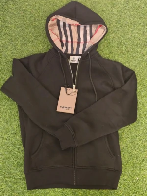 Svart hoodie från Burberry med dragkedja - Svart hoodie från Burberry med klassiska Burberry-ränder i huvan och guldfärgad dragkedja. Hoodien har fickor framtill och justerbar huva med snören. Perfekt för dig som vill ha en stilren och exklusiv look.