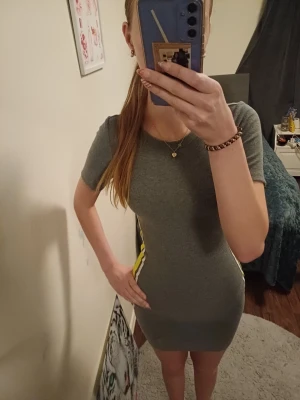 Grå tight kortklänning med ränder - Säljer en grå tight kortklänning med korta ärmar och coola vita och gula ränder längs sidorna. Klänningen har en sportig vibe och är gjord i stretchigt material som sitter snyggt på kroppen. Perfekt för dig som gillar stretchiga kläningar, skriv om ninhat frågor, priset går att diskutera storlek XS men passar även S 