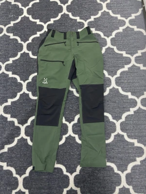 Gröna friluftsbyxor från Haglöfs - Snygga gröna friluftsbyxor från Haglöfs med svarta förstärkningar på knän och bak. Byxorna har dragkedjeförsedda fickor framtill och bälteshällor. Perfekta för utomhusaktiviteter och äventyr. Materialet är slitstarkt och flexibelt.