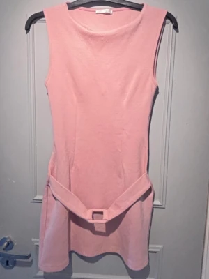 Ljusrosa ärmlös klänning med bälte - Säljer en söt ljusrosa kortklänning i mjukt tyg. Klänningen är ärmlös och har ett matchande bälte i midjan med fyrkantigt spänne. Enkel och stilren design som passar perfekt till sommaren.
