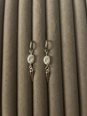 Silvriga örhängen med pärldetalj - Säljer ett par stilrena örhängen i silverfärg med små vita pärlor och spetsiga metallhängen. Örhängena har en modern design med runda pärlor och konformade detaljer som ger en edgy touch. Perfekta för dig som gillar minimalistiska accessoarer.