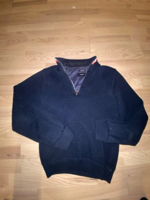 Mörkblå stickad tröja Holebrook - Mörkblå stickad tröja från Holebrook med halv dragkedja och hög krage. Tröjan har vindtätt innerfoder och ribbade muddar vid ärmslut och nederkant. Perfekt för kyliga dagar och snygg till jeans.