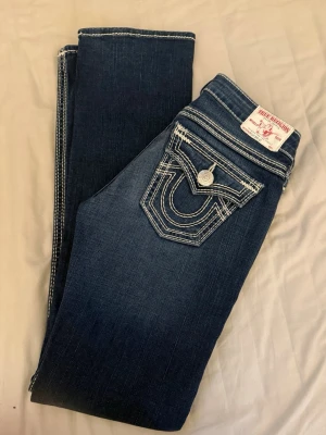 True Religion jeans - Säljer ett par mörkblå jeans från True Religion med kontrasterande vita sömmar och ikonisk fickdetalj bak. Klassisk femficksmodell med knappgylf och tydlig logga på bakfickan. Snygg tvätt och raka ben, perfekta till en avslappnad streetstil. Vintage. Storlek W26, helt nya i skicket 