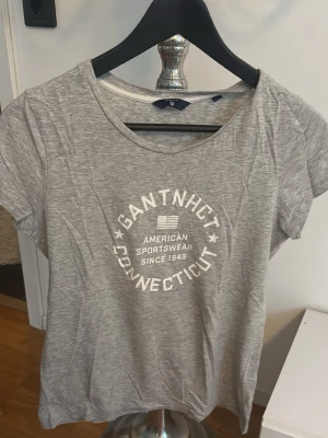 Grå Gant t-shirt med tryck - Snygg grå t-shirt från Gant med vitt tryck framtill där det står 'GANT NHCT CONNECTICUT' och 'AMERICAN SPORTSWEAR SINCE 1949'. Klassisk rund halsringning och korta ärmar. Perfekt till jeans eller shorts.