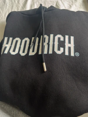 Svart hoodie från Hoodrich - Säljer en svart hoodie från Hoodrich med stor vit logga framtill och mindre tryck på fickan. Klassisk huva med snören och magficka. Materialet känns mjukt och bekvämt, perfekt för en avslappnad streetwear-look.