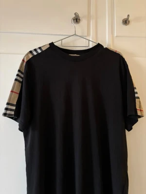 Svart Burberry t-shirt med rutiga ärmar - Snygg svart t-shirt från Burberry med klassiska beige, röda och svarta rutiga detaljer på axlarna. T-shirten har korta ärmar och en relaxed passform. Perfekt för dig som vill ha en stilren men ändå unik look.