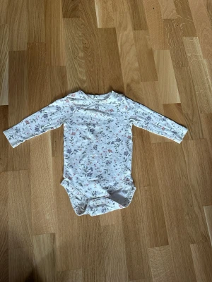 Blommig långärmad body från Newbie - Söt långärmad body från Newbie i vitt med blommigt mönster i rosa, lila och grönt. Bodyn har tryckknappar vid axeln och i grenen för enkel påklädning. Tillverkad i mjukt material som passar perfekt för små barn.