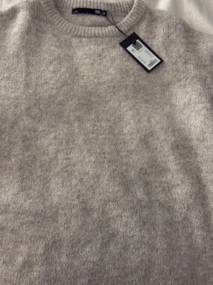 Beige stickad tröja från 157 - Mysig beige stickad tröja från 157 med rund halsringning och ribbade muddar. Tröjan har en enkel och stilren design som passar perfekt till lager-på-lager och är lätt att matcha med olika outfits.