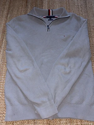 Grå halvzip tröja Tommy Hilfiger - Snygg grå halvzip tröja från Tommy Hilfiger i ekologisk bomull. Tröjan har ribbade muddar, hög krage med dragkedja och diskret logga på bröstet. Perfekt till jeans eller chinos för en clean och stilren look.