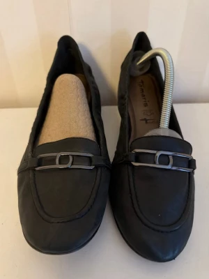 Svarta loafers från Tamaris - Snygga svarta loafers från Tamaris i mjukt läder med låg klack och dekorativ metallspänne framtill. Klassisk design som passar till många olika stilar. Skorna har en rundad tå och diskreta sömmar längs kanterna.