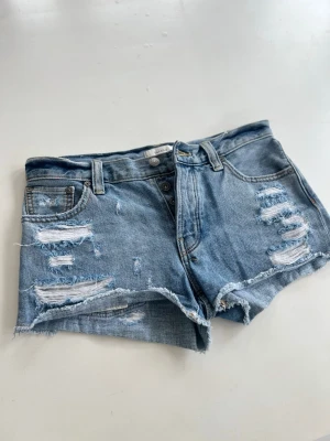 Blå slitna jeansshorts med knappar - Säljer ett par blå jeansshorts med slitna detaljer och råa kanter. Shortsen har knappgylf, bälteshällor och fem klassiska fickor. Perfekta för sommaren och ger en avslappnad vibe.