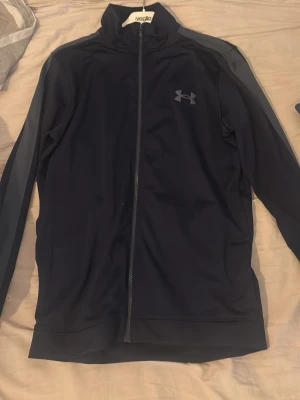 Svart träningsjacka från Under Armour - Snygg svart träningsjacka från Under Armour med hel dragkedja framtill och hög krage. Jackan har diskret logga på bröstet och är tillverkad i ett stretchigt funktionsmaterial som passar perfekt till träning eller uppvärmning. Byxor till hoodien ingår