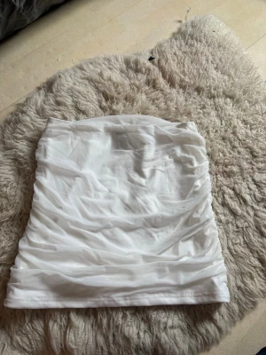 Vit kortkjol med rynkade detaljer - Säljer en vit kortkjol med snygga rynkade detaljer. Kjolen har en tight passform och är gjord i ett stretchigt syntetmaterial som sitter skönt på kroppen. Perfekt för dig som gillar en enkel men trendig look.