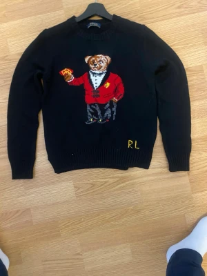 Svart stickad tröja Polo Ralph Lauren - Stickad svart tröja från Polo Ralph Lauren med ikonisk björn i röd kavaj och RL-brodyr framtill. Tröjan har rund halsringning och ribbade muddar vid ärmslut och nederkant. Perfekt för dig som gillar klassiska och lekfulla detaljer. Inte äkta. Är lite för små så den funkar också som xs