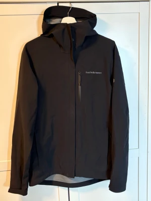 Svart vindjacka Peak Performance - Snygg svart vindjacka från Peak Performance med huva och dragkedja framtill. Jackan har en bröstficka med dragkedja och diskret logga på bröstet. Tillverkad i ett lätt och slitstarkt material som står emot vind och regn. Perfekt för aktiva dagar utomhus.