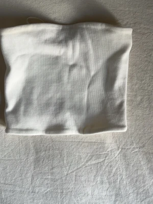 Vit ribbad bandeau tubtopp - Säljer en enkel och stilren vit bandeau tubtopp med ribbad struktur. Perfekt att matcha med  jeans eller kjol för en clean look. Toppen är ärmlös och har en mjuk, stretchig passform. Tröjan är i st S men är ganska liten hade sagt att den passar xs