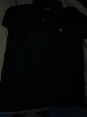 Svart pikétröja från Lyle & Scott - Klassisk svart pikétröja från Lyle & Scott med krage och knappar framtill. Tröjan har diskret logga på bröstet och är tillverkad i ett pikématerial som känns både stilrent och bekvämt.