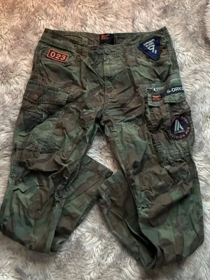 Camo cargobyxor från Superdry - Säljer ett par grymma cargobyxor från Superdry i camomönstrad grön färg. Byxorna har flera coola patchar och detaljer, stora fickor på sidorna och dragkedja. Storlek 33W, 32L men de sitter snyggt även om du har mindre storlek 🖤