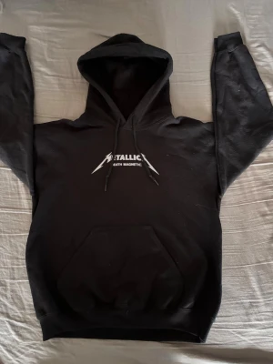 Svart Metallica hoodie med tryck - Säljer en svart hoodie från Metallica med 'Death Magnetic'-tryck på bröstet och ett stort grafiskt tryck på ryggen. Hoodien har huva med dragsko och en stor magficka. Perfekt för dig som gillar bandmerch och vill ha en skön, stilren look. Storleken: S