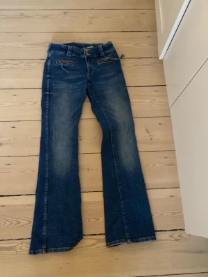 Blå bootcut jeans med dragkedjor - Säljer ett par blå bootcut jeans med snygga slitningar och dragkedjedetaljer framtill. Jeansen har klassisk femficksdesign och utsvängda ben. Perfekta för en avslappnad och trendig look.