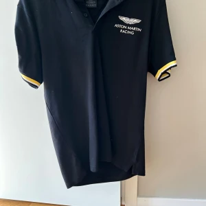 Svart Aston Martin Racing t-shirt - Snygg svart t-shirt från Aston Martin Racing med vit logga på bröstet och tryck på ärmen. T-shirten har korta ärmar med vita och gula detaljer vid ärmsluten. Perfekt för dig som gillar motorsport och stilren design. Helt oanvänd.