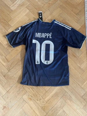 Real Madrid matchtröja Mbappé blå - Snygg blå Real Madrid matchtröja från Adidas med Mbappé och nummer 10 på ryggen. Tröjan har vita detaljer, klubbmärke och sponsortryck på bröstet samt klassiska Adidas-ränder på axlarna. Tillverkad i lätt och ventilerande material, perfekt för fotbollsträning eller match.