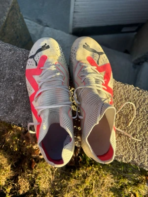 Puma vita fotbollsskor med neonröd detalj - Säljer ett par vita fotbollsskor från Puma med högt skaft och snörning. Skorna har en tydlig neonröd zigzag-detalj på sidorna och svarta Puma-loggor på tårna. Ovandelen är i syntetmaterial med ventilerande hål för extra komfort. Skorna har inga problem, de funkar super bra 