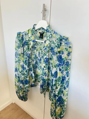 Blommig blus från Lindex med volanger - Blus från Lindex i ett färgstarkt blommigt mönster i blått, grönt och gult. Modellen har volangdetaljer vid axlarna, knytband i halsen och långa ärmar med resår vid ärmsluten. Tunn och luftig kvalitet, perfekt för en trendig look.