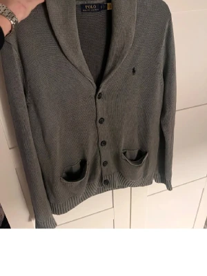 Grå kofta från Polo Ralph Lauren - Snygg grå kofta från Polo Ralph Lauren med sjalkrage och knäppning framtill. Två fickor och diskret broderad logga på bröstet. Perfekt att slänga på sig över en t-shirt för en chill men ändå stilren look. Storlek S men sitter som M