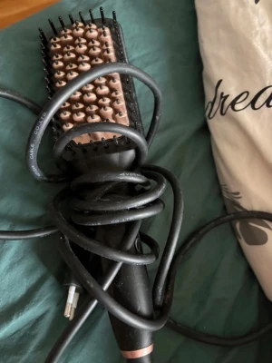 Elektrisk värmeborste för hår - En elektrisk värmeborste med svart handtag och borsthuvud i roséguld. Borsten har täta piggar och sladd för elanslutning. Perfekt för att styla och räta ut håret snabbt och smidigt.