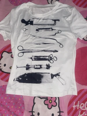 Vit t-shirt med kirurgiska verktyg - Säljer en vit t-shirt med tryck av olika kirurgiska verktyg i svart. T-shirten har rund hals och korta ärmar. Perfekt för dig som gillar unika och edgy motiv. Materialet känns mjukt och bekvämt mot huden.